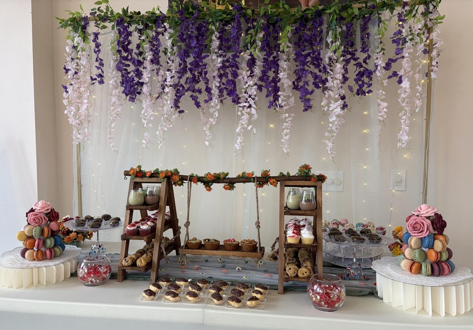 Full event dessert table styled with cascading purple and blush wisteria backdrop, macaron towers, cupcakes and cookies
